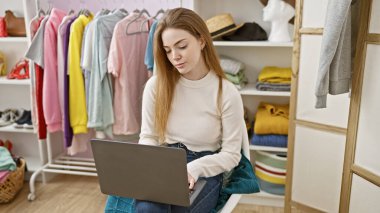 Genç sarışın bir kadın, renkli bir gardıropta dizüstü bilgisayar kullanarak içeride oturuyor, kıyafetlerle çevrili..