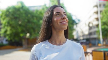 Şehir parkında gülümseyen bir kadın. Arka planda yemyeşil alanlar ve şehir binaları var.