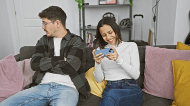 Oturma odasında yanında oturan sıkkın bir adamla akıllı telefon kullanan bir kadın..