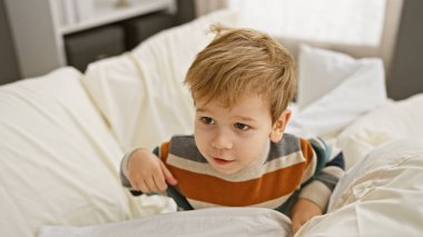 Sarışın küçük çocuk, beyaz bir yatakta otururken meraklı görünüyor..