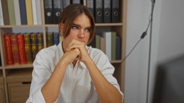Genç, esmer bir kadın ofis ortamında arka planda kitaplıklarla gündüz vakti derin düşüncelere dalıyor..