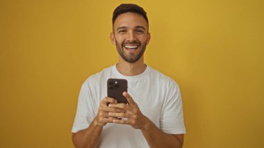 Zeki bir telefon tutan, parlak sarı bir arkaplan önünde gülümseyen, kapalı bir ortamda beyaz tişört giyen, yakışıklı İspanyol bir genç..