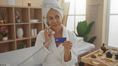 Spa ortamında orta yaşlı bir kadın telefonda konuşuyor ve elinde bir kredi kartı var. Etrafı sağlık ürünleriyle çevrili ve evde dinlendirici bir atmosferi var.