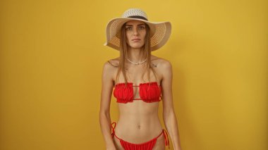 Kırmızı bikinili ve güneş şapkalı güzel bir beyaz kadın izole edilmiş sarı arka plan duvarına karşı poz veriyor..
