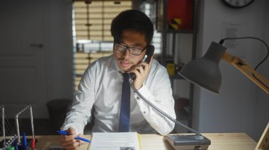 Gömlekli ve kravatlı genç bir Asyalı telefonda konuşuyor. Ofis malzemeleri ve belgelerle çevrili..