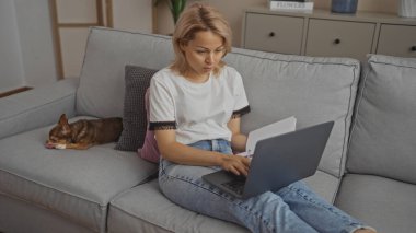 Genç sarışın bir kadın laptopuyla oturma odasındaki gri bir kanepede çalışıyor. Köpeği yanında dinleniyor, rahat ve rahat bir ortam yaratıyor..