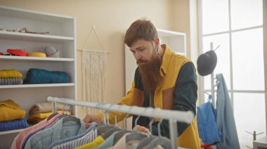 Sakallı adam güneşli, modern bir terzide renkli tekstil rafları olan elbiseleri inceliyor..