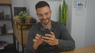 Sakallı ve gözlüklü yakışıklı, genç, İspanyol bir adam ofis ortamında bitki ve raflarla süslenmiş bir akıllı telefon kullanıyor..