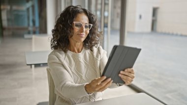 Profesyonel İspanyol bir kadın modern ofis ortamında bir tablet kullanıyor. Uzmanlık ve nişan duygusunu yansıtıyor..