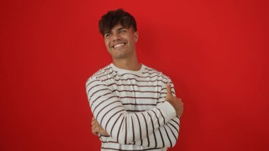 Kolları çaprazlanmış yakışıklı İspanyol bir adam izole edilmiş kırmızı duvarlara gülümsüyor ve çizgili bir gömlek giyiyor..