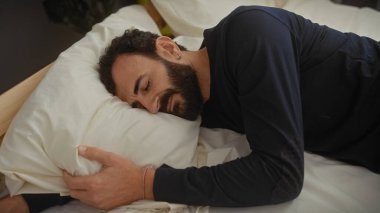 Siyah gömlekli, sakallı, orta yaşlı bir adam yatak odasında beyaz bir yastığın üzerinde huzur ve rahatlık içinde yatıyor..