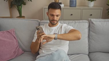 Oturma odasındaki kanepede oturan yakışıklı, sakallı genç bir adam, evde telefon tutarken akıllı hareketlerini kontrol ediyor..