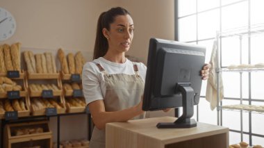 Woman working and looking surprised at computer screen in a bakery filled with bread, showing bustling indoor shop setting with bright natural lighting clipart