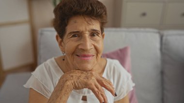 Kısa saçlı, yaşlı bir İspanyol kadın sıcak bir gülümsemeyle oturma odasında oturur ve olgun ve kıdemli güzelliğini evinde gösterir..