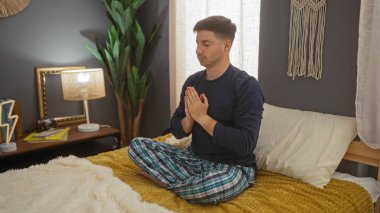 Genç bir İspanyol adam meditasyonda elleri dua pozisyonunda rahat bir yatak odasında iç mekan bitkileri ve hafif ışıklandırmalı bir yatakta oturur..