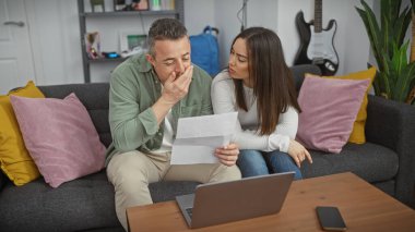Endişeli bir kadın, arka planda dizüstü bilgisayarı ve gitarıyla rahat bir oturma odasında gazete tutan bir adamı rahatlatır..