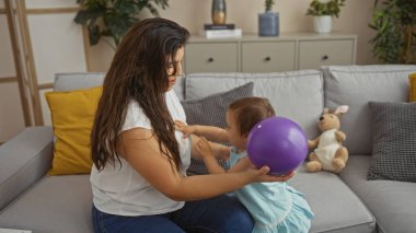 Anne ve kızı, oturma odasında mor bir balon ve arka planda oyuncak bir kanguru ile etkileşime girerek hassas bir anı paylaşıyorlar.