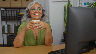 Gri saçlı bir kadın kulaklıkla müzik dinliyor ve ofis ortamında elinde bardak tutuyor.