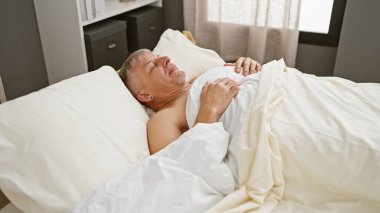 Gri saçlı bir adam minimalist bir yatak odasında huzur içinde uyuyor. Huzur ve istirahat tasvir ediyor..