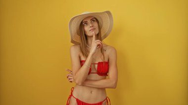 Kırmızı bikinili ve güneş şapkalı çekici sarışın kadın izole edilmiş sarı arka plana nazikçe poz veriyor..