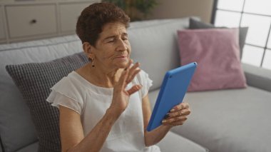 Mavi tabletle sohbet eden kısa saçlı yaşlı İspanyol kadın sıcak bir oturma odasında oturuyor, el sallıyor ve gülümsüyor..