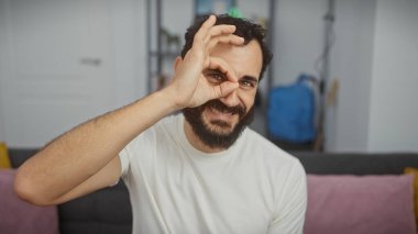 Gülümseyen sakallı bir adam modern bir oturma odasında hoş bir jest yapıyor..