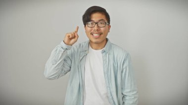 Gözlüklü gülümseyen Asyalı genç adam parmak kaldırarak bir noktaya parmak basıyor, beyaz izole arka plana karşı gelişigüzel giyiniyor..