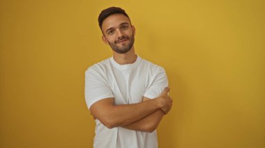 Sakallı ve çapraz kollu genç adam izole edilmiş sarı arka plana gülümsüyor.