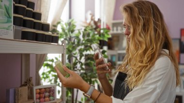 Sarışın bir kadın akıllı telefonuyla bir ürünün fotoğrafını çekiyor. Ev dekorasyonları ve yeni sabun malzemeleriyle çevrili.