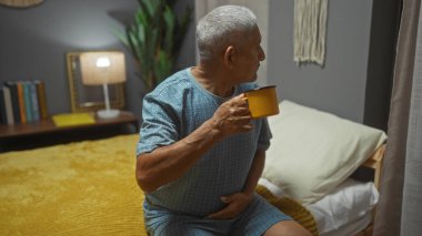 Beyaz saçlı yaşlı İspanyol bir adam, sıcak, kapalı bir yatak odası ortamında bir yatakta otururken sarı bir kupa tutuyor..