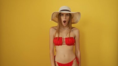 Kırmızı bikinili ve güneş şapkalı güzel sarışın bir kadın izole edilmiş sarı bir arkaplanda hayret verici bir ifadeyle duruyor..