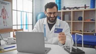 Sakallı bir doktor, parlak bir klinikte bilgisayarının başında otururken kredi kartını inceliyor..