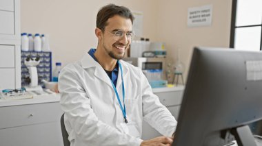 Laboratuvar önlüklü, sakallı, gülümseyen genç İspanyol adam laboratuvar ortamında bilgisayar kullanıyor.