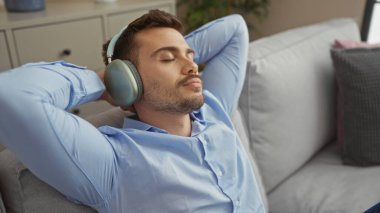 Yakışıklı, sakallı, genç, İspanyol bir adam oturma odasında kulaklıklarla dinleniyor ve bir dairede sakin bir ev ortamı sergiliyor..