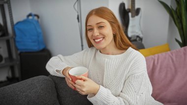 Beyaz kazaklı, gülen kızıl saçlı kadın elinde bardakla, rahat ve sade bir ortam sunuyor..