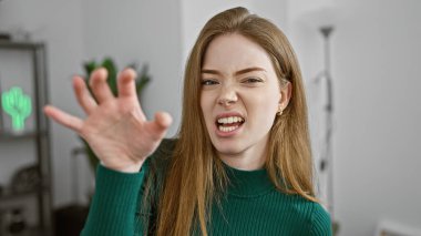 Yeşil kazaklı genç bir beyaz kadın, modern bir ev içinde hoşnutsuzluk göstererek elleriyle el kol hareketi yapıyor..
