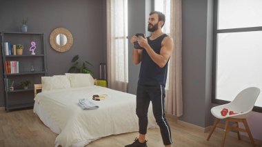 Modern bir yatak odasında dambılla egzersiz yapan yakışıklı bir adam, evde sağlıklı bir yaşam tarzını yansıtıyor..