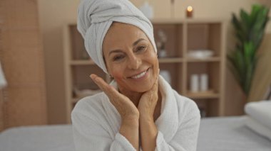 Rahatlatıcı bir spa ortamında havlu başlığı ve bornozla gülümseyen bir kadın kapalı bir salon ortamında olgun güzellik ve sağlığını sergiliyor..