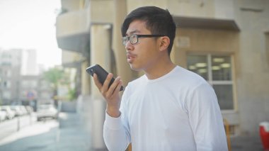 Gözlüklü genç bir Asyalı şehir sokaklarında akıllı telefon kullanıyor, şehir yaşam tarzı ve teknolojisini somutlaştırıyor..