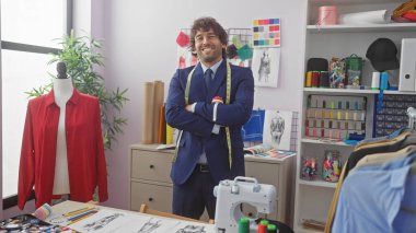 Kolları çapraz duran, sakallı, gülümseyen İspanyol bir adam. Renkli bir terzi dükkanında. Etrafı dikiş malzemeleri ve çizimlerle çevrili.