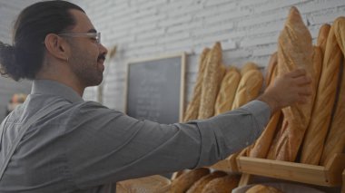 Bıyıklı İspanyol bir adam arka planda çeşitli taze somunlar sergilenen sıcak bir pastanenin içinde ekmek seçiyor..