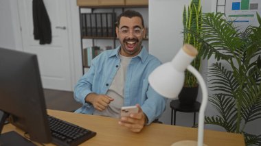 Ofiste akıllı telefon kullanırken bıyıklı, gülümseyen genç İspanyol bir adam.