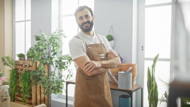 Sakallı, önlüklü, kendine güvenen bir adam, parlak bir çiçekçide kollarını kavuşturuyor..