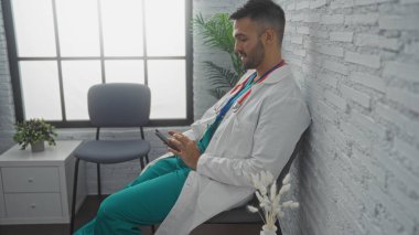 Klinik bekleme odasında sakallı, beyaz önlüklü, akıllı telefon kullanan genç bir İspanyol..