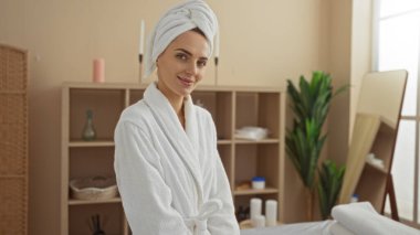 Beyaz bornoz ve havlu giymiş, kaplıcada sakin bir atmosferde gülümseyen çekici sarışın kadın..