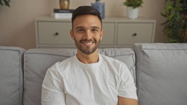 Sakallı yakışıklı bir İspanyol genç modern bir oturma odasında oturuyor, gülümsüyor, beyaz bir gömlek giyiyor ve parlak ve rahat bir dairede doğrudan kameraya bakıyor..