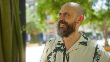 Sakallı ve kel kafalı yakışıklı İspanyol bir adam. Arka planda yeşil ve şehir binaları olan bir parkta gülümsüyor.