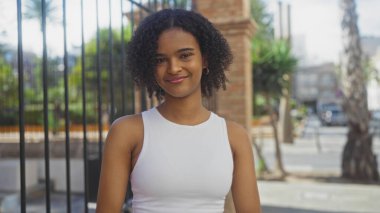 Kıvırcık saçlı, güzel, Afro-Amerikalı bir kadın şehir merkezinde güneşli bir sokakta kendine güvenerek gülümsüyor..