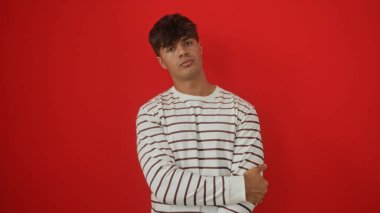 Beyaz çizgili tişörtlü genç bir İspanyol adam kollarını kırmızı bir arka planda kavuşturmuş, izole edilmiş bir portrede çekici ve kendinden emin bir adamı tasvir ediyor..
