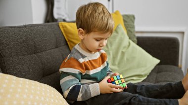 Sarışın çocuk gri bir kanepede rahat bir şekilde otururken renkli bir bulmaca küpünü çözmeye odaklanmış..